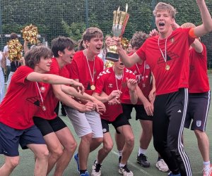 na boisku zewnętrznym na pierwszym planie 7 cieszących się z wygranej chłopców w czerwonych koszulkach ze złotymi medalami, jeden z nich trzyma puchar. W tle cheerleader'ki oraz chearleader'rzy dopingujący zwycięzców trzymają złote pompony