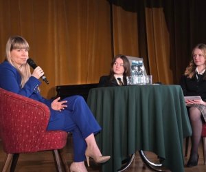 na szkolnej scenie znajduje się stolik z zielonym nakryciem, na którym stoją 3 butelki wody oraz 3 szklanki. Przy stoliku na pierwszym planie siedzi kobieta, Monika Horna-Cieślak, ubrana w niebieski garnitur przemawia do mikrofonu, natomiast na drugim planie siedzą 2 dziewczyny ubrane w szkolne mundurki, które z zaciekawieniem słuchają przemowy kobiety, Rzecznika Praw Dziecka