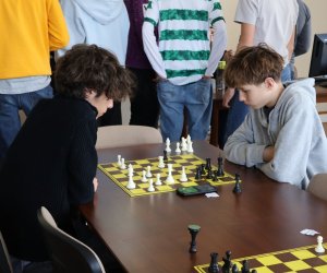 w centralnej części zdjęcia 2 skupionych uczniów rozgrywa partię szachów. W tle znajduje się duże grono chłopców, które jest zwrócone tyłem do kamery